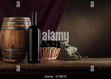 Vino rosso bottiglia, canna e carrello con uve su una tavola in legno rustico, panno rosso su sfondo, degustazione di vino ancora vita Foto Stock