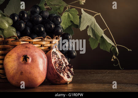 Le uve in un cestello e melograni con foglie di vite su un tavolo di legno, frutta ancora vita Foto Stock
