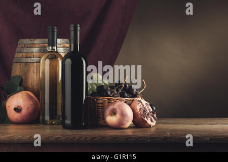 Le bottiglie di vino, canna e cesto di frutta su una tavola in legno rustico, panno rosso su sfondo, still life Foto Stock