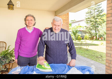 La stiratura Senior nel patio della sua casa mentre un altro uomo affettuosamente trattiene la sua mano sulla sua spalla Foto Stock