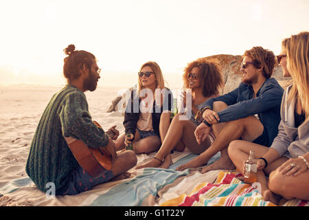 Gruppo di amici in un momento di relax a un sunset beach party con un giovane uomo di suonare una chitarra. I giovani aventi un party sulla spiaggia. Foto Stock