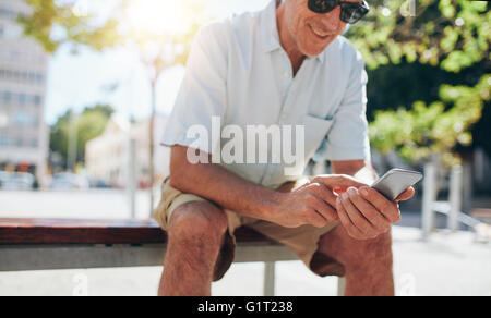 Ritratto di tagliate di sorridere uomo maturo usando il cellulare mentre seduti all'aperto su un banco in città Foto Stock