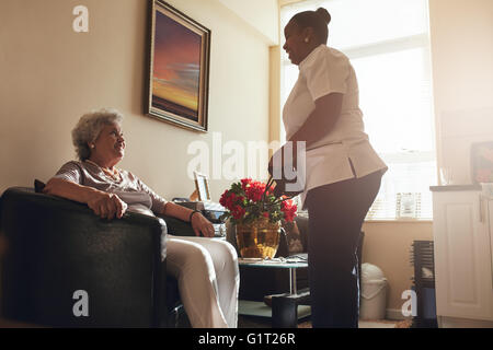 Senior donna seduta su una sedia a casa con il caregiver femmina in attesa. Femmina visita infermiere paziente senior per il controllo di blo Foto Stock