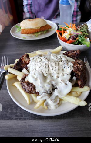 Una piastra di manzo con salsa di funghi e i chip con una ciotola di insalata greca e schnitzel di pollo Burger in background Foto Stock