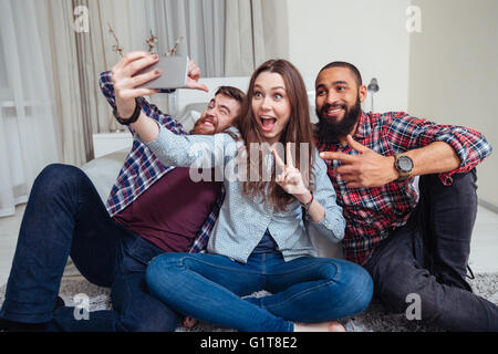 Gruppo di felice giovani amici seduti e tenendo selfie a casa Foto Stock