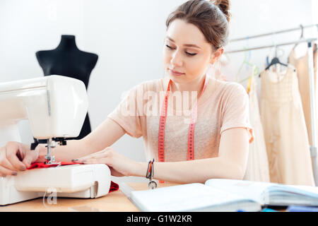 Concentrato bella giovane donna sarta cuce sulla macchina per cucire e lettura delle note in notebook Foto Stock