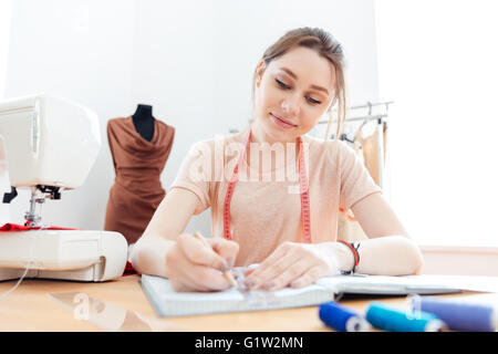 Sorridente piuttosto giovane donna sarta disegno pattents in notebook al lavoro Foto Stock