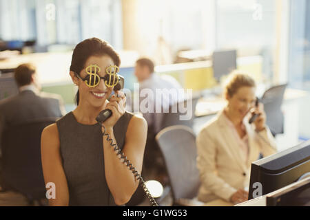 Ritratto di sorridere imprenditrice che indossa il simbolo del dollaro occhiali da sole rispondendo telefono in ufficio Foto Stock