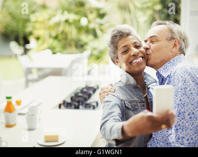 Affettuosa Coppia matura kissing tenendo selfie in cucina Foto Stock