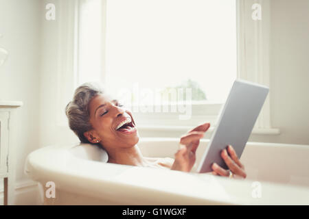 Ridendo donna matura con tavoletta digitale nella vasca da bagno Foto Stock