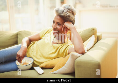 Confortevole donna matura di guardare la TV e mangiare popcorn sul divano Foto Stock