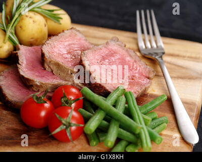 Foto di bistecca per cena con spesse fette di lombata di pomodori ciliegini, fagiolini e patate su una tavola di legno. Foto Stock