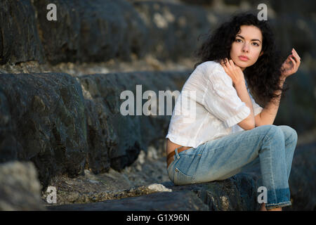 Un giovane attraente slim brunette donna ragazza nel profondo del pensiero, seduti da sé da soli , jeans blu, da solo da se stessa all'aperto al di fuori in una serata calda. Regno Unito Foto Stock