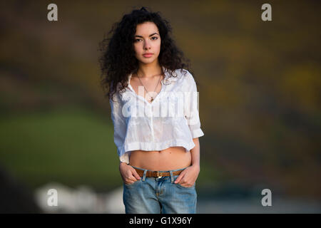 Un giovane attraente slim brunette donna ragazza adolescente con lunghe parentesi ondulato marrone capelli in piedi all'aperto al di fuori in una serata calda indossano jeans e ritagliata camicetta bianca, rivolta in avanti, REGNO UNITO Foto Stock