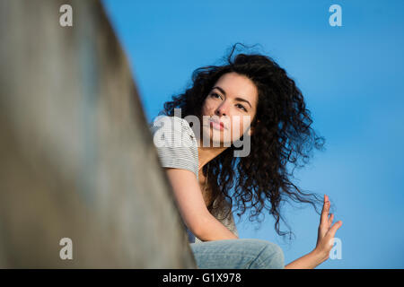 Un giovane attraente slim brunette donna ragazza con lunghi ricci selvatici capelli castani, all'aperto al di fuori su un cielo blu calda serata. Regno Unito Foto Stock