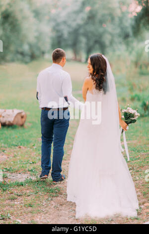 Bello lo sposo e la sposa in bianco velo tornare indietro a piedi tenendo le mani sullo sfondo verde della foresta Foto Stock