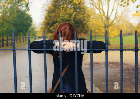 Un triste donna è in appoggio la sua testa su un cancello nel parco in autunno Foto Stock