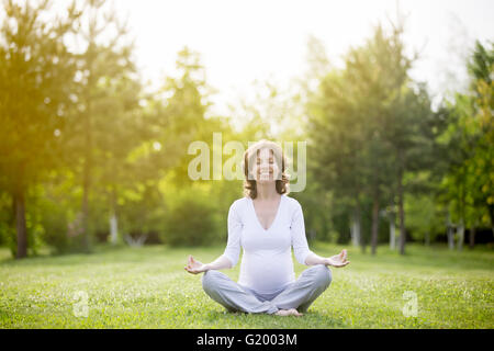 Ritratto di giovane modello incinte lavorano nel parco. Sorridente futura mamma aspetta bambino seduto a gambe incrociate e la meditazione Foto Stock