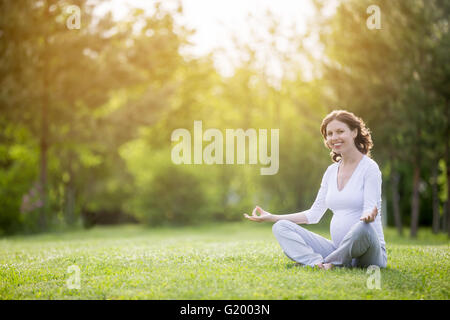 Ritratto di giovane modello incinte lavorano nel parco. Sorridente futura mamma aspetta bambino seduto a gambe incrociate sulla meditazione Foto Stock