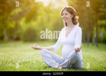 Ritratto di giovane modello incinte lavorano nel parco. Sorridente futura mamma seduta con le gambe incrociate sulla sessione di meditazione sulla natura Foto Stock