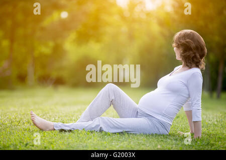 Felice giovani modello incinta seduta sul prato e guardando il sole che tramonta nel parco. Sorridente futura mamma aspetta baby Foto Stock