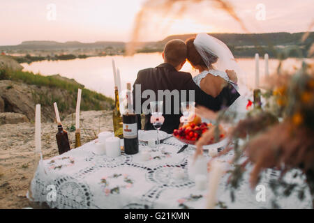 I giovani sposi siiting torna al lago durante la serata romantica con candele sulla spiaggia di sabbia Foto Stock