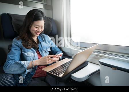 Donna asiatica sorridente a smartphone con computer portatile sul treno, spazio copia sulla finestra Foto Stock