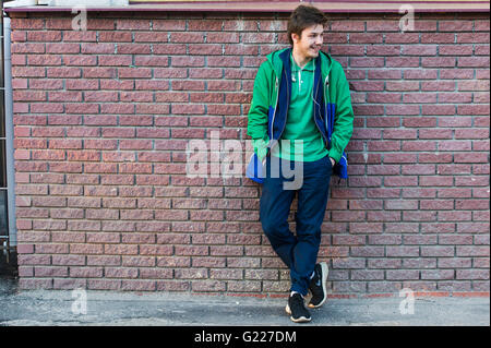 Comune di giovane uomo verde che indossa la t-shirt camicia e jeans contro in rosso di un muro di mattoni cercando lateralmente Foto Stock