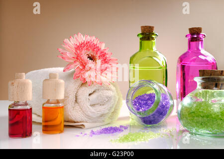 Oli e sali da bagno bianco sul tavolo di vetro. Decorata con fiori e asciugamano marrone con gradiente dello sfondo. Composizione orizzontale Foto Stock