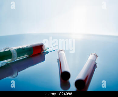 Siringa e fiale con il prelievo di campioni di sangue oltre il tavolo di vetro e lo sfondo è blu. Composizione orizzontale. Vista posteriore. Foto Stock