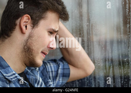 Vista laterale di un uomo triste guardando attraverso la finestrella quasi gridando in un giorno di pioggia Foto Stock