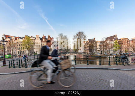 Persone in bicicletta attraverso strade di città su un bel giorno di estate in Amsterdam, Paesi Bassi Foto Stock