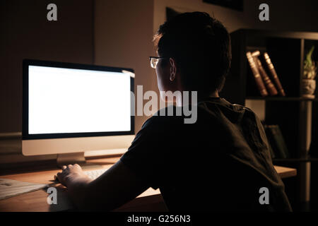 Vista posteriore del giovane serio con gli occhiali seduto e lavorando con schermo vuoto computer a casa durante le ore notturne Foto Stock