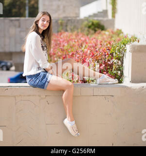 Moda giovane studente ragazza seduta su un muro di mattoni. Outdoor ritratto della ragazza sorridente in pantaloncini e maglietta bianca Foto Stock