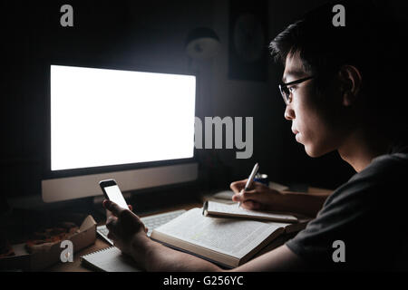 Focalizzato asian giovane uomo con schermo vuoto del telefono cellulare e computer e scrivere note in Blocco note in ore notturne Foto Stock