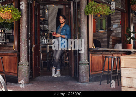 A piena lunghezza Ritratto di rilassata giovane maschio in piedi all'ingresso di una caffetteria e di un caffè. Elegante giovane a un cafe ent Foto Stock