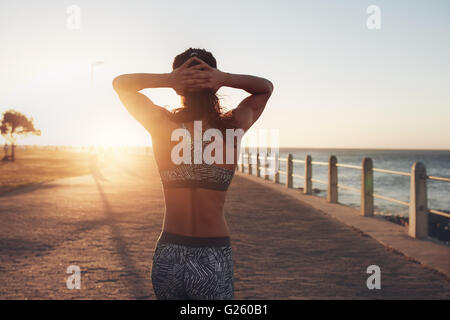 Vista posteriore immagine della donna rilassata nell'abbigliamento sportivo a piedi lungo il mare con le mani sulla sua testa. Sportive facendo una passeggiata da th Foto Stock