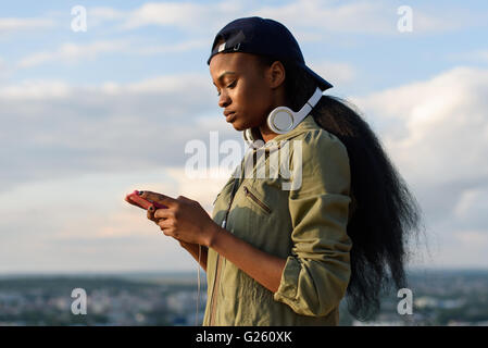 Bella African American Girl ascoltare musica e gode. Sorridente giovane donna nera su sfocata sullo sfondo della città Foto Stock