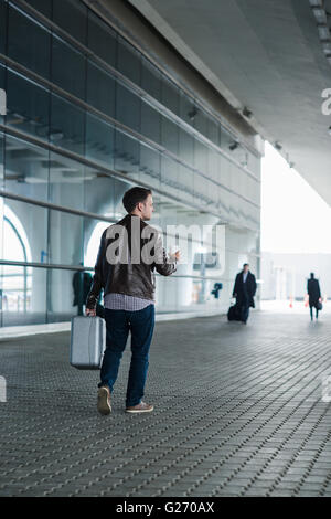 Ritratto di profilo di un giovane uomo che cammina con la valigia e la tazza di caffè vicino al terminal aeroportuale Foto Stock