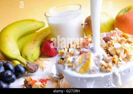 Flusso di latte di cadere in una ciotola di cereali e frutta su un tavolo con frutta e bicchiere di latte sullo sfondo Foto Stock