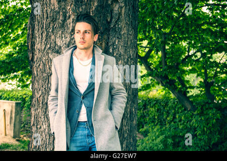 Bel giovane uomo appoggiato contro l'albero, guardando la telecamera, in una giornata di sole indossando un leggero cappotto grigio Foto Stock