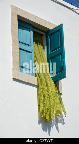 Un verde lime tendina scorre fuori da una finestra aperta in una cornice quadrata con persiane blu in un bianco lavato reso parete, Grecia Foto Stock