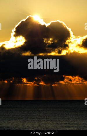 I raggi di sole esplodere attraverso le nuvole in fasci di luce arancione che si tuffano nel mare Foto Stock
