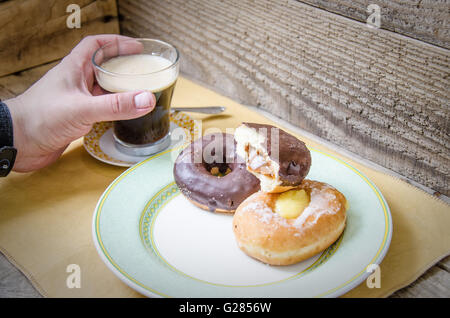 Mano afferrando una tazza di caffè e deliziose ciambelle su una piastra con pallet Sfondo legno Foto Stock