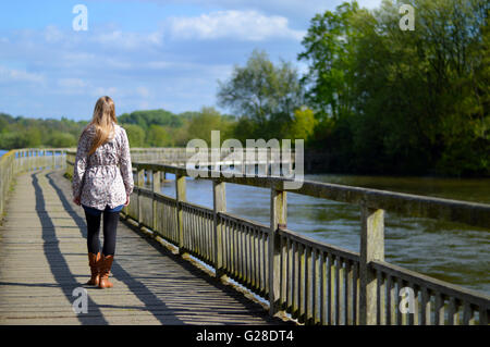 Una donna bionda camminando lungo un ponte di legno sul fiume in una giornata di sole con il blu del cielo. Foto Stock