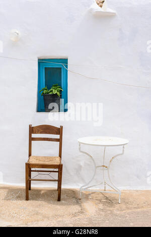 Sedia singola e un piccolo tavolo sotto una finestra blu al di fuori di una casa dipinta di bianco a Sifnos, Grecia Foto Stock