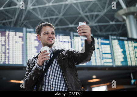 Sorridente giovane viaggiatore uomo con un caffè presso l'aeroporto rende selfie nella parte anteriore della scheda calendario Foto Stock