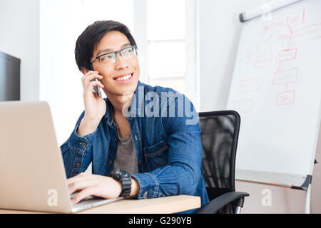 Sorridente asian giovane imprenditore con laptop parlando al telefono cellulare in ufficio Foto Stock