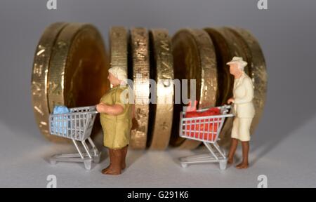 Due figurine in miniatura donna anziana spingendo un supermercato carrello della spesa tra british una libbra di monete. Foto Stock