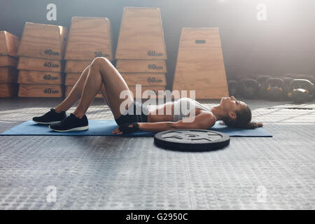 Vista laterale colpo di donna fitness in appoggio sul tappeto di esercizio con una pesante piastra di peso sul pavimento. Atleta donna sdraiata sulla schiena a poppa